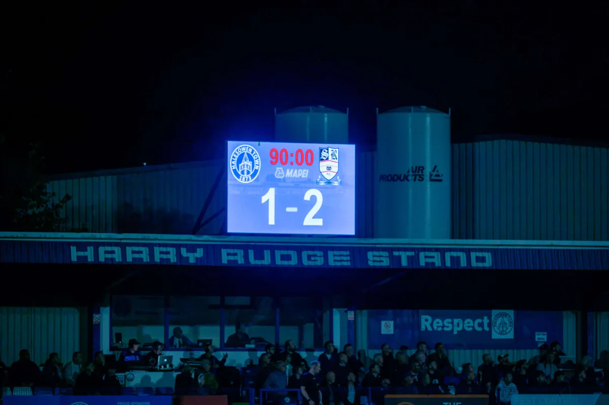The scoreboard at Halesowen Town's ground reading 90:00 and 1-2, above the Harry Rudge Stand with spectators below